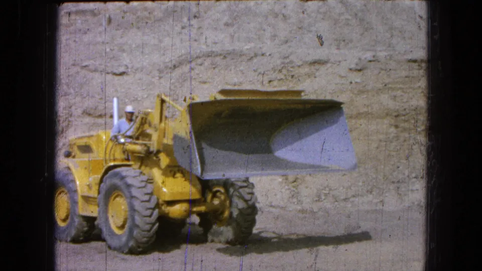 1965 Pueblo, Mexico 8mm frame showing the bucket loader in profile with the raised bucket dominating the worksite view.