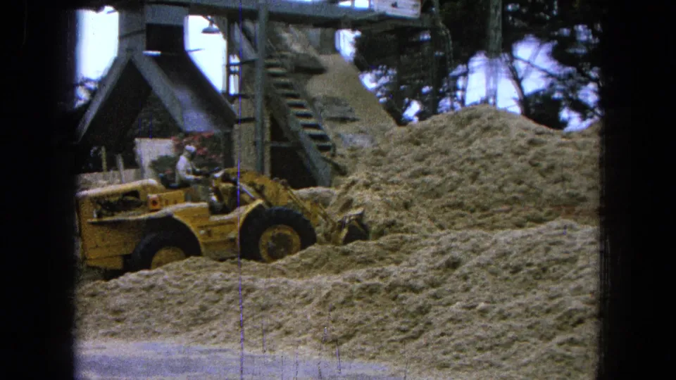 1965 Pueblo, Mexico 8mm frame showing a yellow front-end loader near large pale piles with metal plant structures behind it.