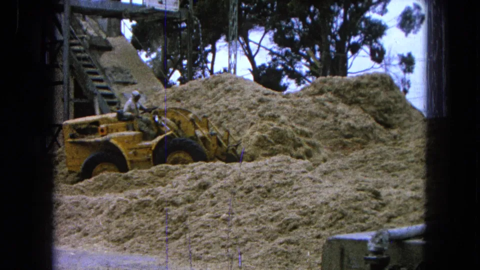 A 1965 Pueblo, Mexico 8mm view shows the loader partly surrounded by pale piles, with a worker at the controls and machinery behind.