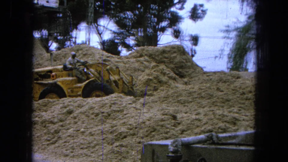 Final 1965 Pueblo, Mexico frame showing the yellow loader working along the pale mound under trees and plant structures.