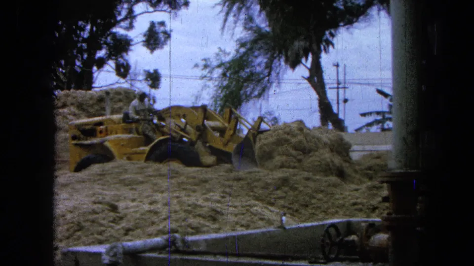 A grainy 8mm view shows the loader close to a sand pile, framed by dark film edges and roadside utility lines in Pueblo, Mexico.