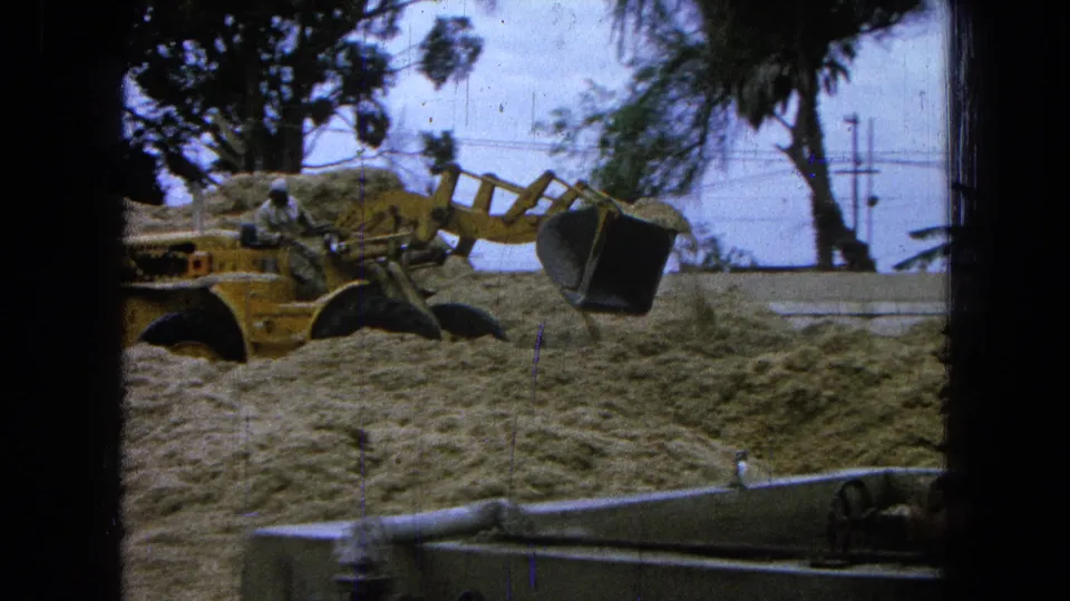 The loader lifts its bucket above the sand in this 1965 Pueblo, Mexico frame, with the worker, palms, utility poles, and foreground piping visible.