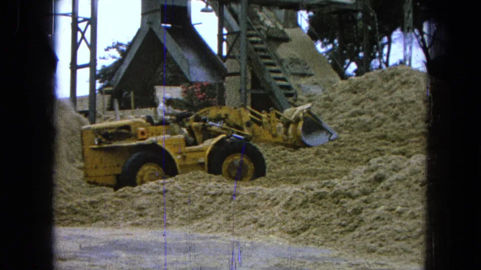 The loader and operator appear below metal framing and a chute, with sand piles filling the foreground in the 1965 worksite scene.
