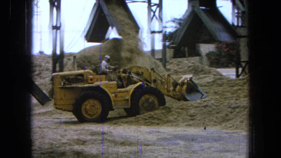 A yellow front-end loader with a hard-hat operator sits near a chute and sand piles in the Pueblo, Mexico construction footage.