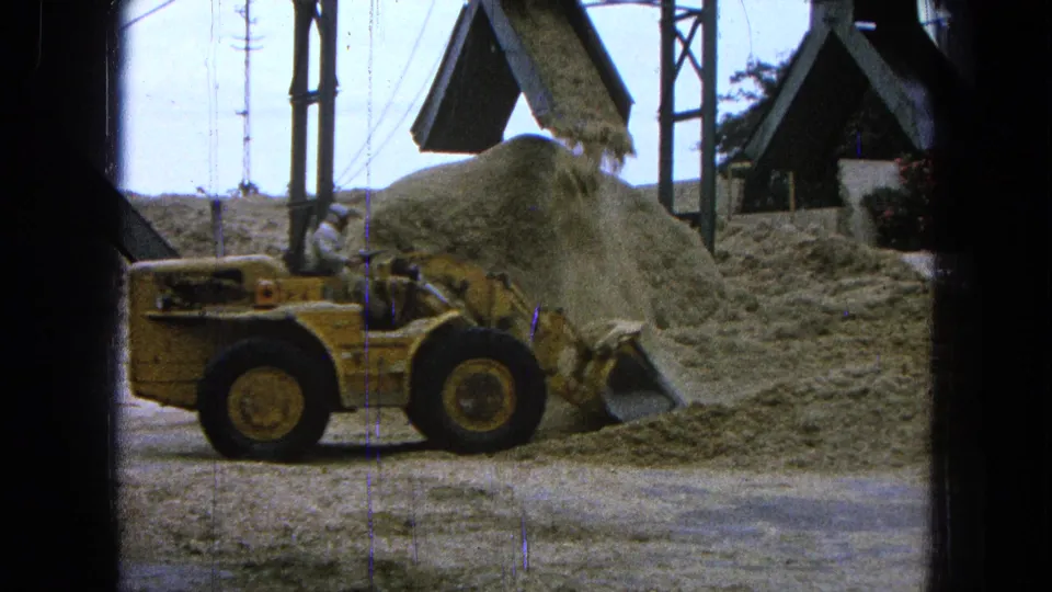 1965 Pueblo, Mexico 8mm frame showing a yellow payloader near a mound as sawdust drops from sawmill equipment.