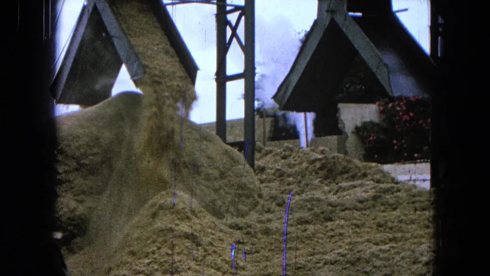 1965 Pueblo, Mexico footage shows aggregate pouring from a conveyor beside small roofed structures and material piles.