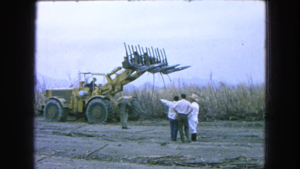 The yellow loader lifts cane in a Pueblo, Mexico field in 1965 while several adults stand nearby observing the work.
