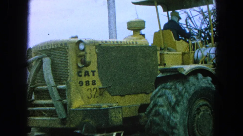 1965 Pueblo, Mexico frame showing the side of a yellow CAT 988 machine, its large tire, and an operator seated above the body.