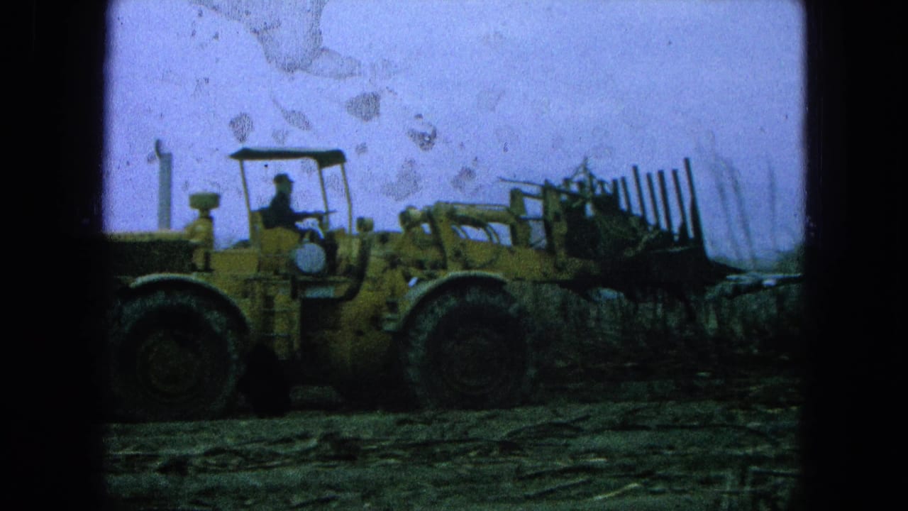Yellow front-end loader with a driver crosses rough field ground under a pale sky.