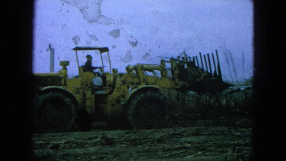 1965 Pueblo Mexico 8mm frame showing a yellow loader in profile with a seated driver and rough field terrain.