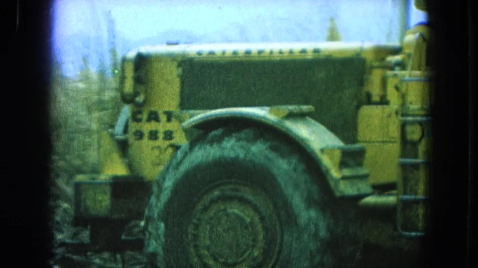 Pueblo Mexico 1965 8mm frame showing the side of a yellow CAT 988 loader as it moves past vegetation.