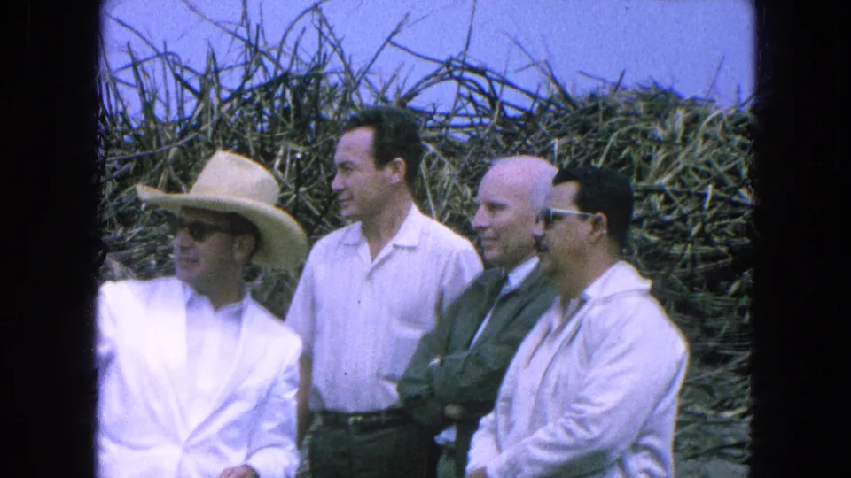 Four adult men in Pueblo, Mexico, 1965, stand at the edge of a harvested field, including one in a straw hat and white suit.
