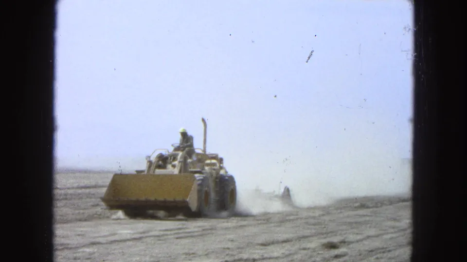 A yellow tractor continues field work in Pueblo, Mexico, 1965, moving across dry soil with a broad dust trail behind it.
