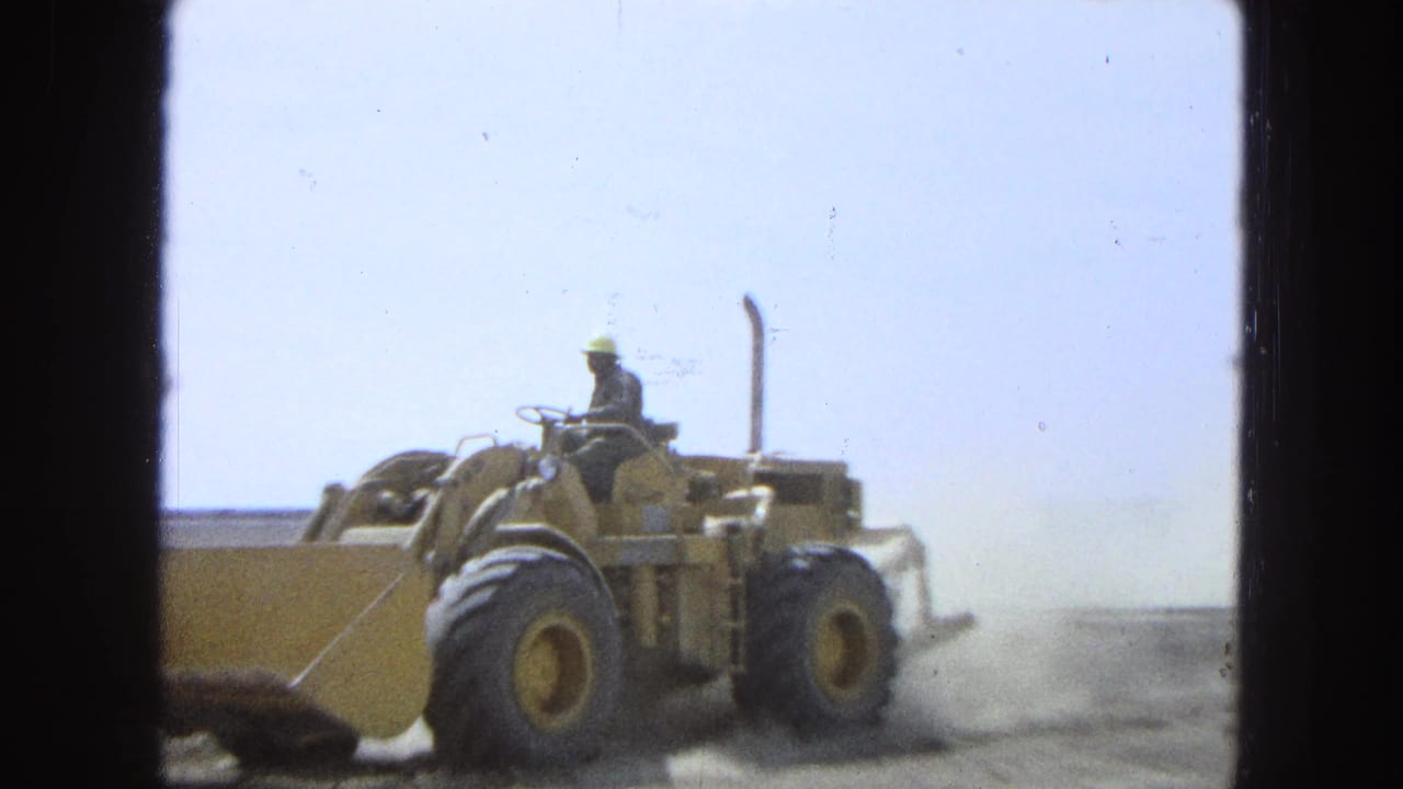 1965 Pueblo, Mexico 8mm frame showing a worker operating a yellow front-end loader across dusty ground under a bright sky.