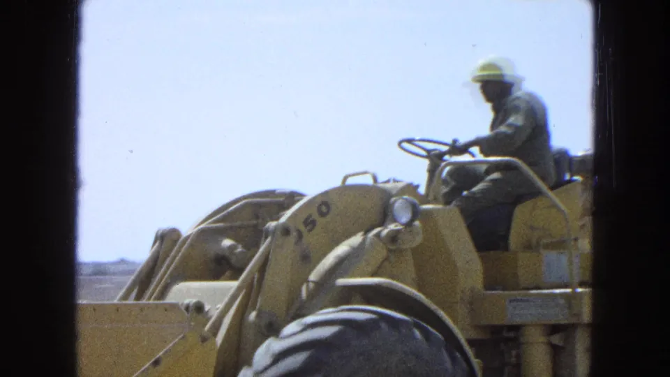 Close 1965 Pueblo, Mexico frame of a yellow front-end loader, with the operator at the wheel and machine markings visible.