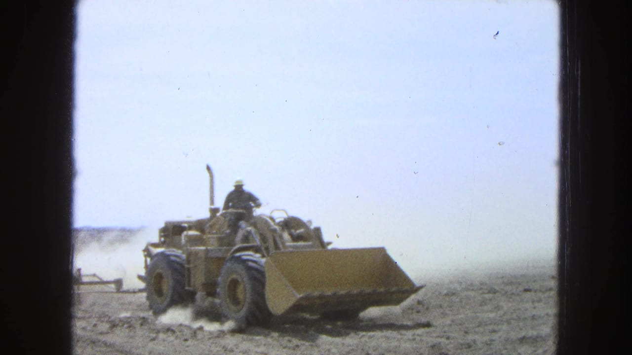 1965 Pueblo, Mexico 8mm frame showing a yellow tractor with an operator pulling equipment through a dry field.