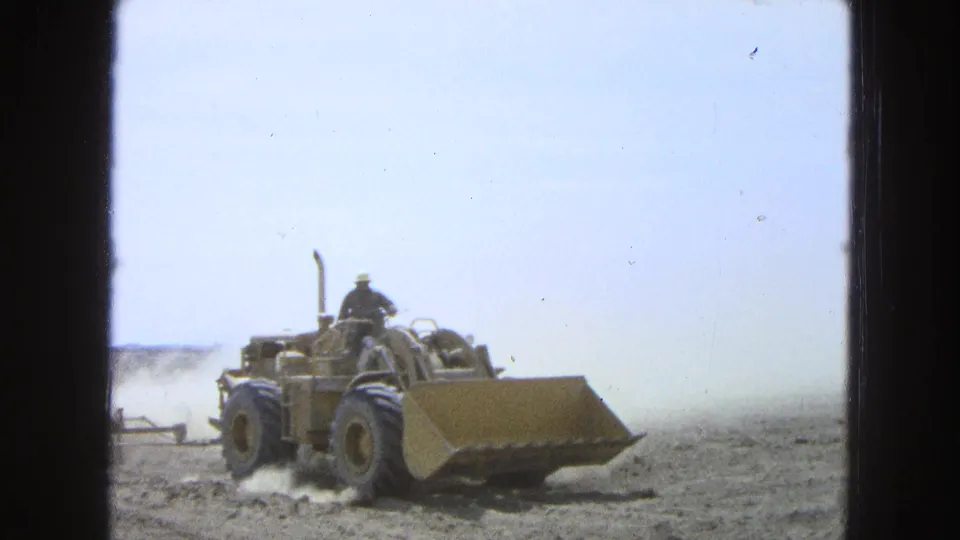 1965 Pueblo, Mexico 8mm frame showing a yellow tractor with an operator pulling equipment through a dry field.