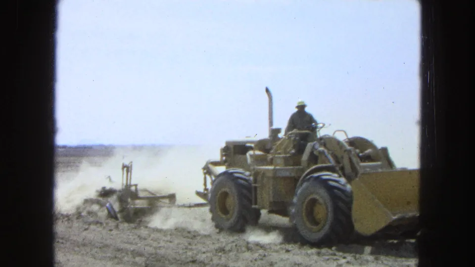 1965 Pueblo, Mexico 8mm frame showing the tractor nearer to camera, with the operator above large tires and dust behind.