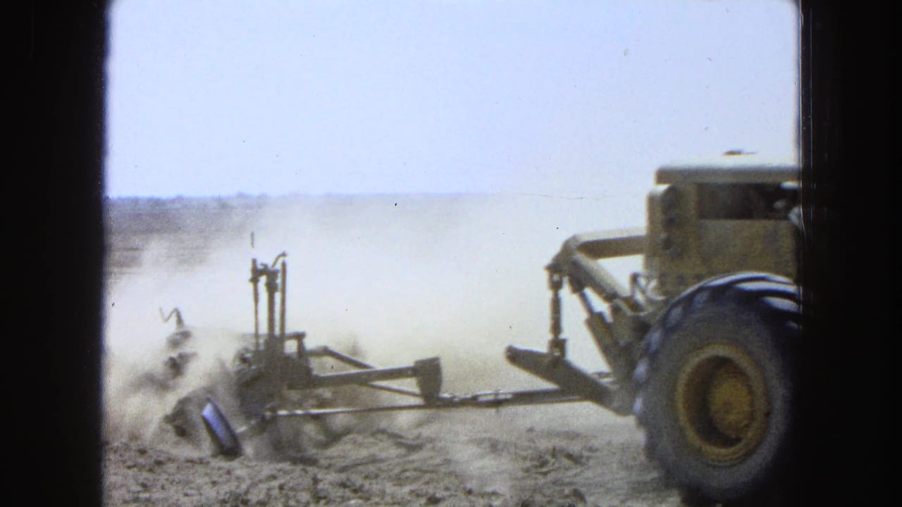 1965 8mm footage from Pueblo, Mexico shows a large tractor and plow moving through a dry field as dust lifts behind the equipment.
