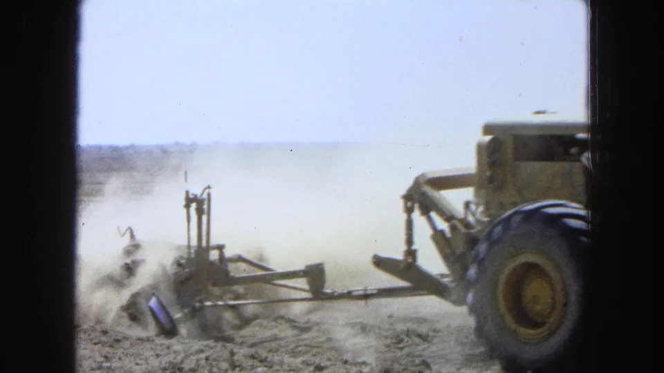 1965 8mm footage from Pueblo, Mexico shows a large tractor and plow moving through a dry field as dust lifts behind the equipment.