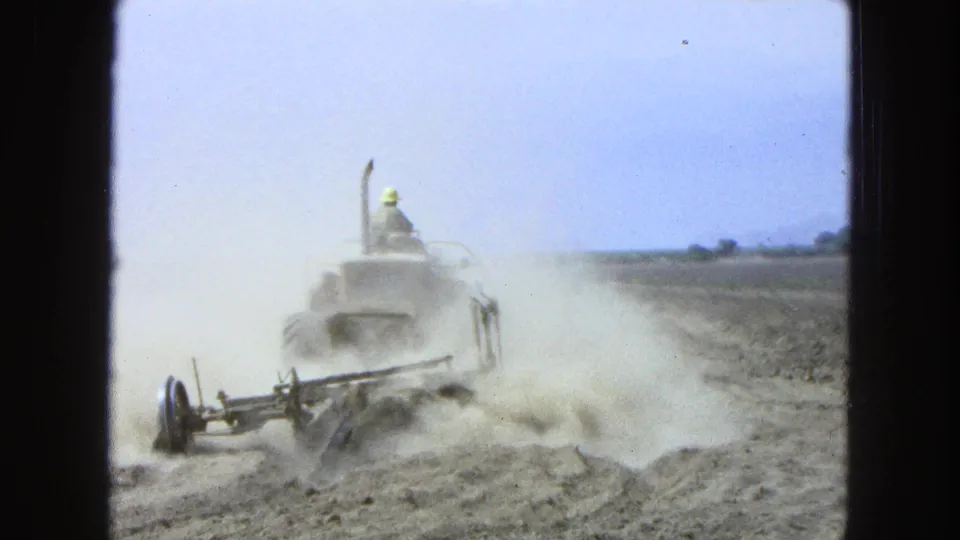 Rear view of a tractor operator in 1965 Pueblo, Mexico guiding a farm machine through a dry field as dust billows outward.