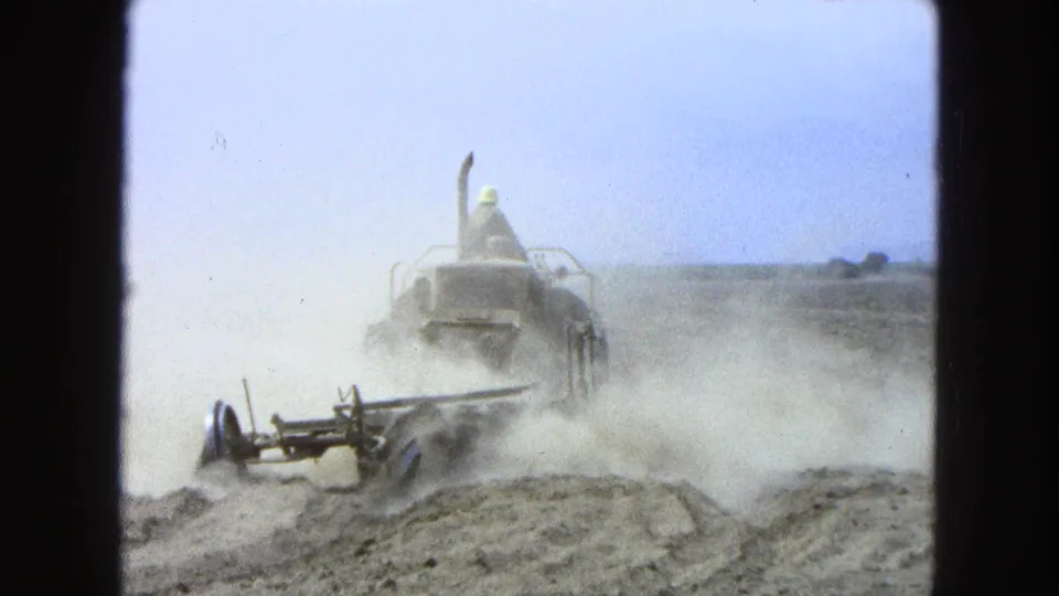 1965 Pueblo, Mexico frame showing the tractor and operator partly veiled by dust during field work on open farmland.
