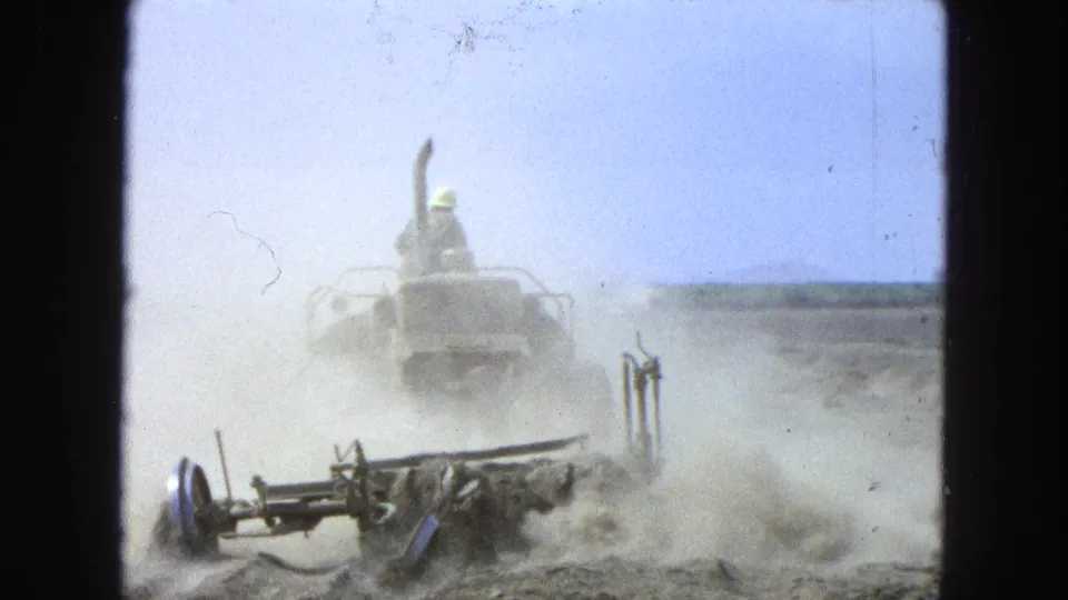 A tractor operator drives forward through dust in a dry field, with plow hardware visible near the camera.