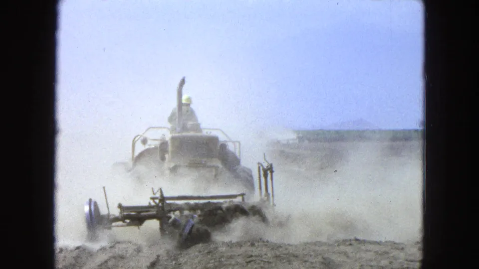 A distant tractor operator drives through dust while plowing a dry field in archival 8mm footage from Pueblo, Mexico.