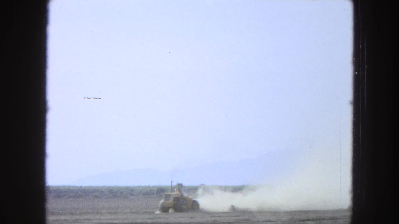 Small construction vehicle crossing a dusty field with a large pale dust cloud behind it.