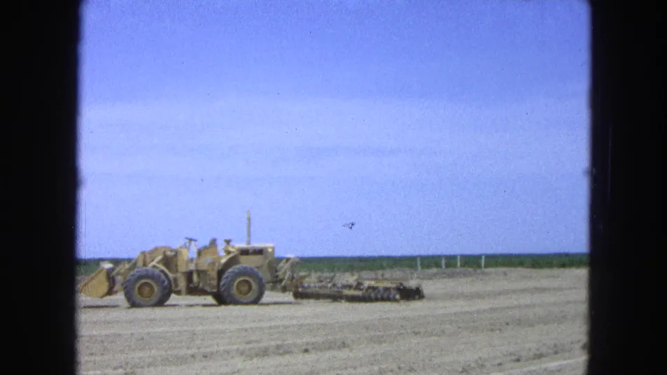 Yellow heavy machinery appears in profile on a dry field in Pueblo, Mexico, with dragged equipment behind it and a low green horizon.