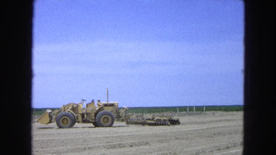 A 1965 8mm view shows yellow earthmoving equipment in a broad dirt field, with sparse horizon detail and visible film-frame borders.