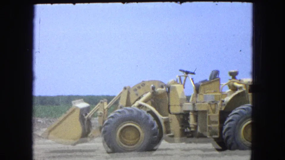 Close 8mm frame of a yellow front-end loader in Pueblo, Mexico, showing large tires, bucket, dust near the ground, and open field beyond.