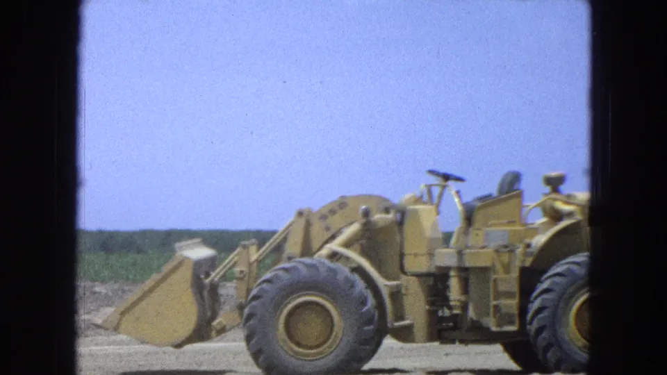 1965 Pueblo, Mexico 8mm frame showing a yellow front-end loader and tractor moving past a dry field with crop rows behind.