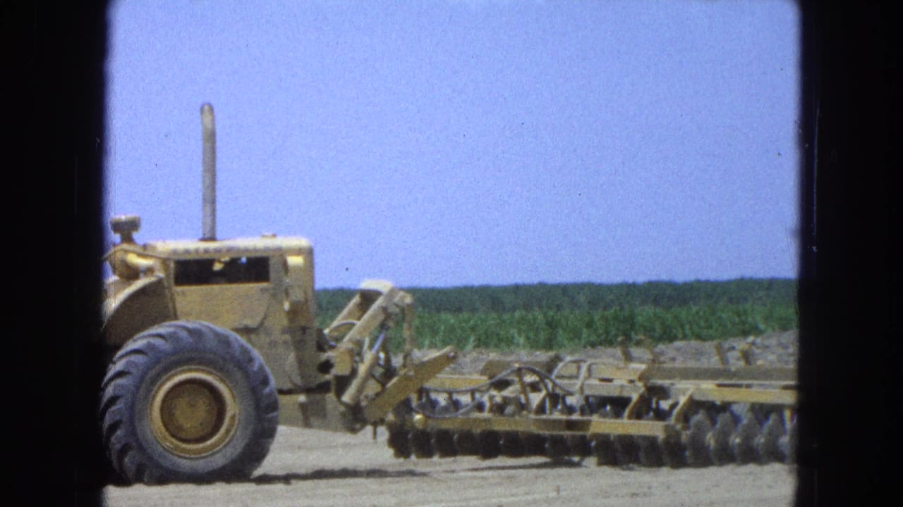 Yellow tractor pulling a disc harrow beside a green field.