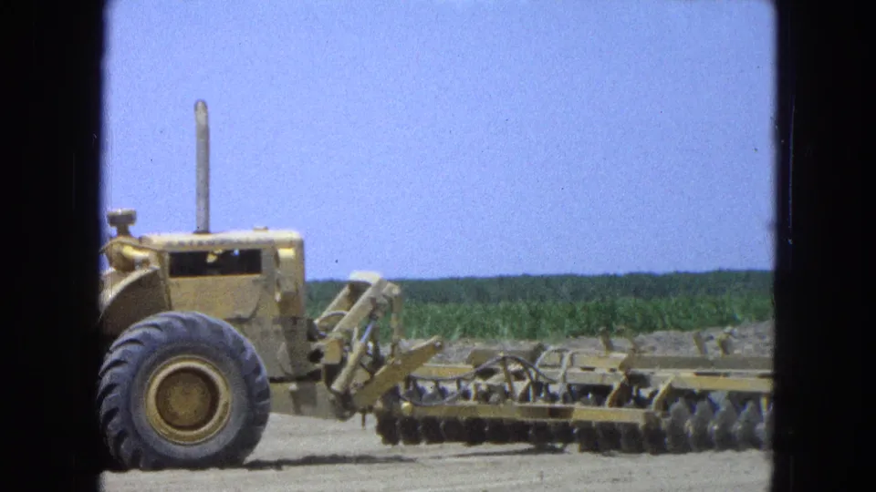 1965 Pueblo, Mexico 8mm frame with a yellow tractor and trailing disc harrow moving along the edge of a cultivated field.