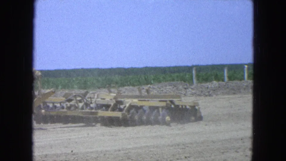 1965 Pueblo, Mexico 8mm frame showing a disc harrow trailing across a dry field, with green crop rows and a low horizon behind.