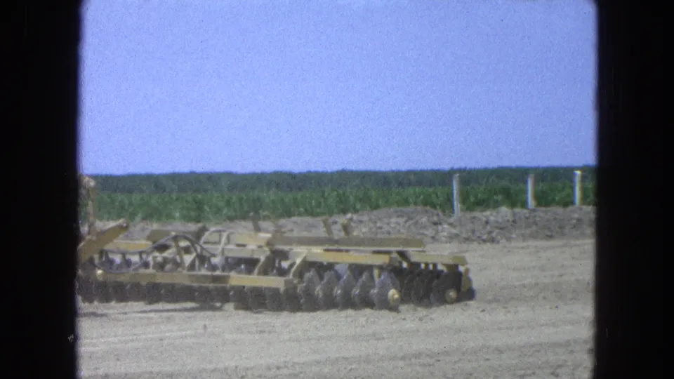 1965 Pueblo, Mexico 8mm frame with a disc harrow continuing across a dry field, seen through dark 8mm contact-sheet borders.
