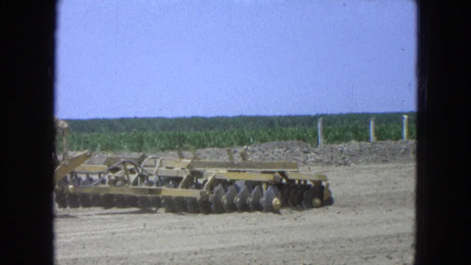 1965 Pueblo, Mexico home-movie frame showing yellow agricultural equipment and disc harrow sections beside a wide prepared field.