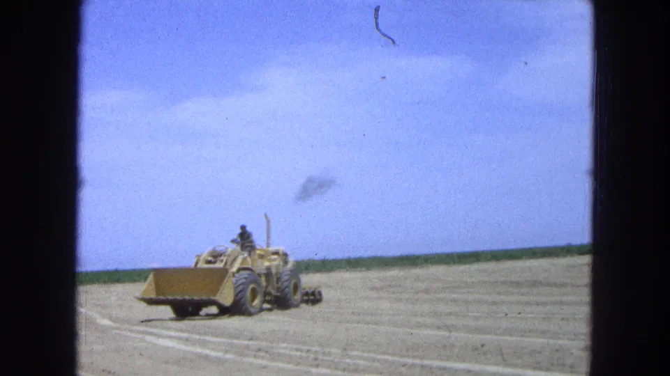 Home-movie frame from 1965 Pueblo, Mexico showing a yellow loader crossing tilled ground with an operator seated above the bucket.