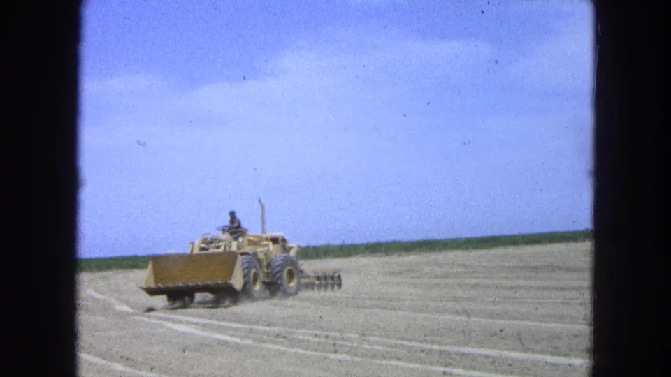 1965 Pueblo, Mexico frame showing a yellow loader and trailing disc harrow working across a broad tilled field under open sky.
