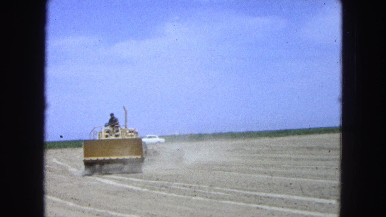 The dozer moves farther across the dry field in 1965 Pueblo, Mexico footage, with dust trailing and a white vehicle far behind.