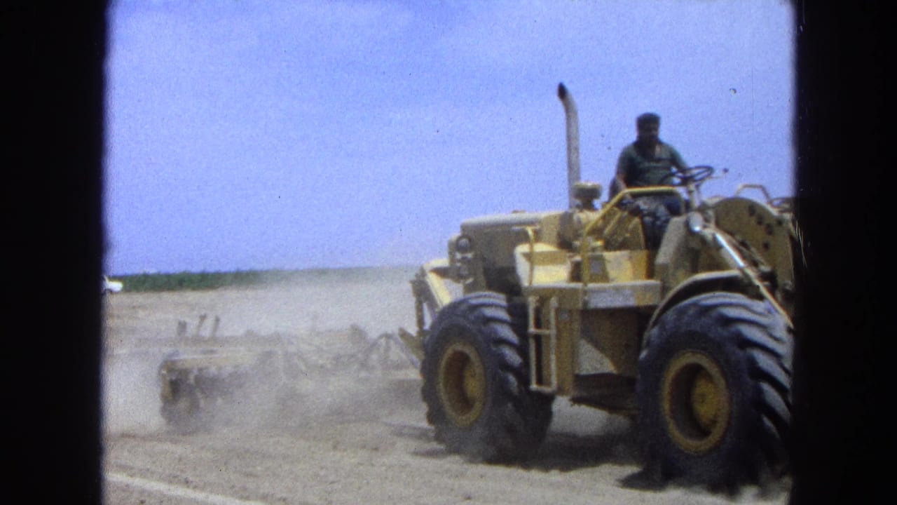 Yellow tractor with a seated operator pulling equipment across dusty farmland.