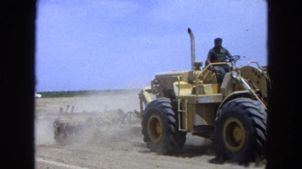 1965 Pueblo, Mexico 8mm frame showing a man on a yellow tractor crossing dry farmland with dust behind the machinery.