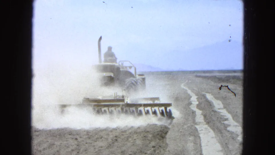 1965 Pueblo, Mexico 8mm frame with a tractor pulling a harrow through heavy dust beside a rough dirt track.