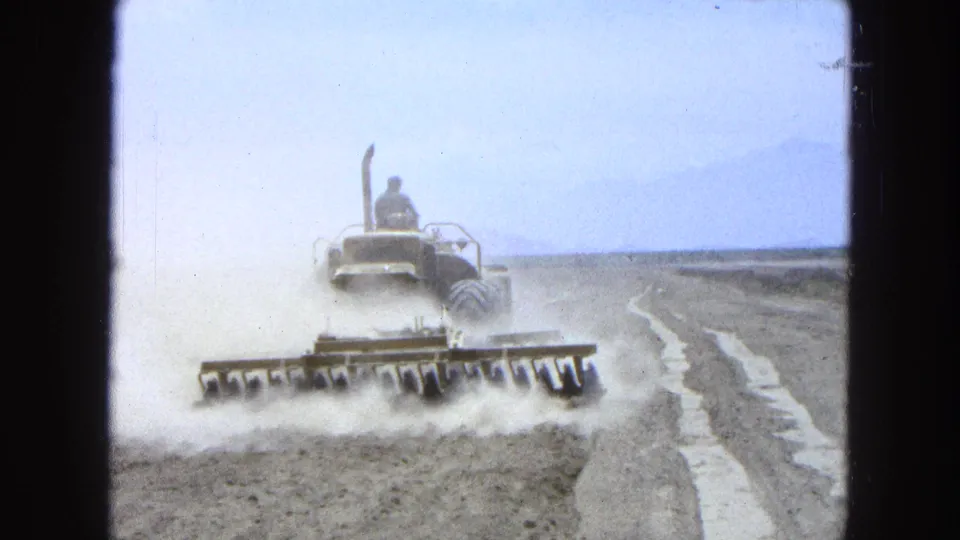 1965 Pueblo, Mexico 8mm frame showing a tractor receding across dry farmland with a wide harrow and dust plume.
