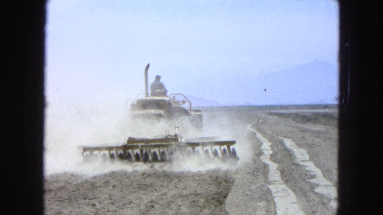 Farmer drives a tractor pulling a disc harrow across a dusty dry field.