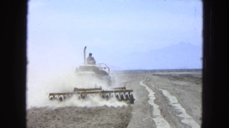 1965 Pueblo Mexico home-movie frame with a tractor-drawn harrow moving along dry ground as dust trails behind it.