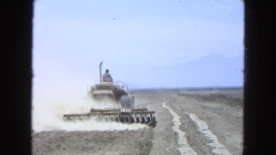 1965 Pueblo Mexico 8mm frame showing the tractor from behind as the harrow stirs dust across a dry agricultural field.