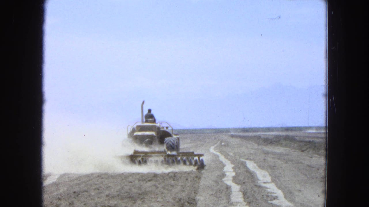 A distant tractor continues across a dry Pueblo, Mexico field in 1965, with dust trailing from the harrow.