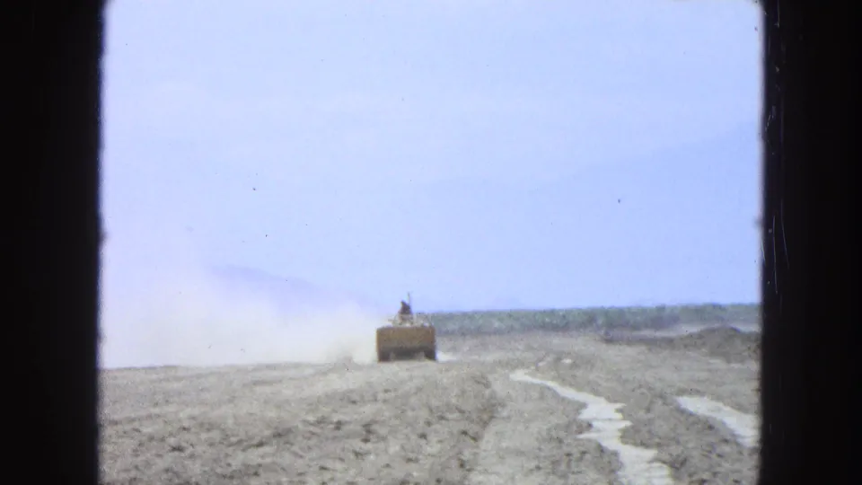 1965 Pueblo, Mexico 8mm frame showing a distant tractor working across dry, pale soil under a light sky.